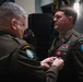 A military award ceremony is taking place in which one U.S. Army officer is pinning a medal onto another officer's uniform. The recipient is wearing a Special Forces Airborne uniform with numerous ribbons, badges, and unit patches. An American flag is visible in the background.