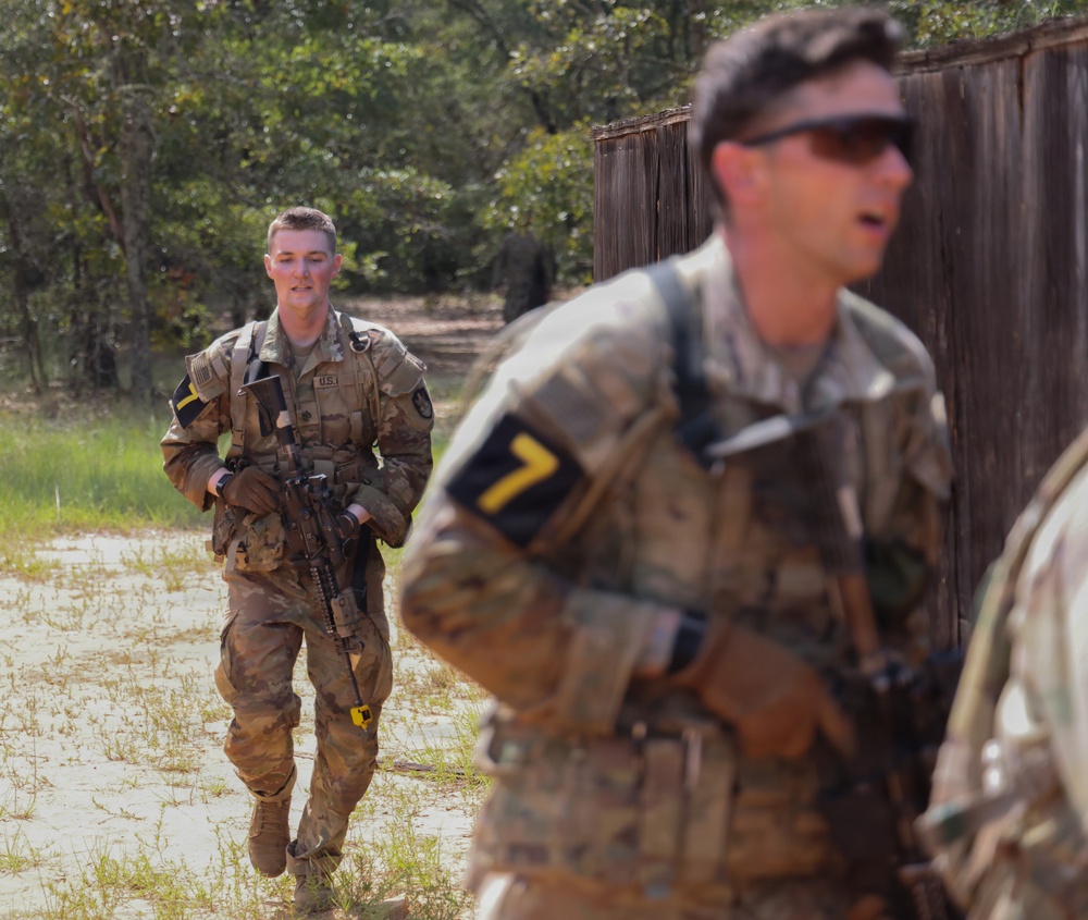 The image shows U.S. Army soldiers in MultiCam (OCP) camouflage uniforms conducting what appears to be a field training exercise. At least two soldiers are clearly visible wearing numbered armbands (number '7') and carrying M4-style rifles. The setting appears to be a training area with sandy terrain, wooden structures, and tree line in the background.