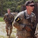 The image shows U.S. Army soldiers in MultiCam (OCP) camouflage uniforms conducting what appears to be a field training exercise. At least two soldiers are clearly visible wearing numbered armbands (number '7') and carrying M4-style rifles. The setting appears to be a training area with sandy terrain, wooden structures, and tree line in the background.
