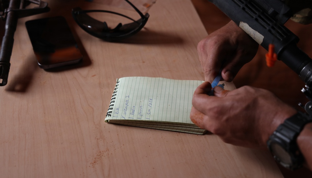 A person is writing in a small spiral-bound notebook on a wooden surface. The notebook contains a handwritten list with partially legible entries. Several items are visible on the table including what appears to be a firearm, sunglasses, and an electronic device.