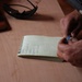 A person is writing in a small spiral-bound notebook on a wooden surface. The notebook contains a handwritten list with partially legible entries. Several items are visible on the table including what appears to be a firearm, sunglasses, and an electronic device.