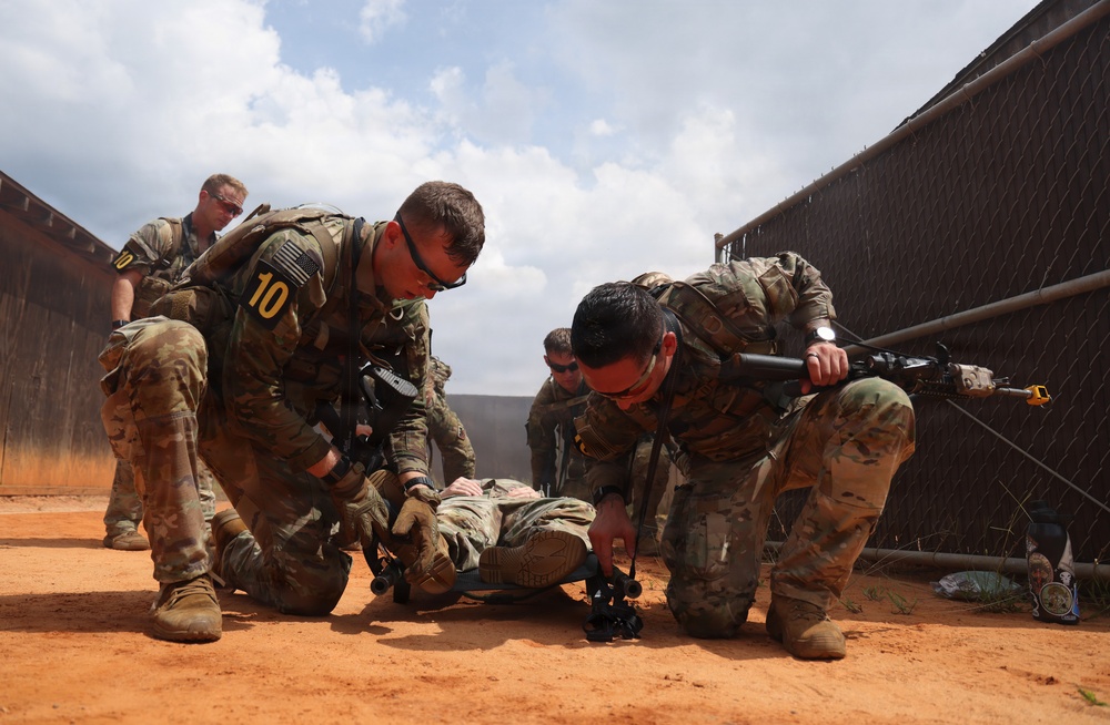 Military personnel in camouflage uniforms are conducting what appears to be a combat casualty care or tactical combat training exercise. Several soldiers are attending to a person lying on red dirt ground, practicing casualty extraction or medical treatment procedures. The setting appears to be a training facility enclosed by chain-link and solid fencing.