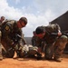 Military personnel in camouflage uniforms are conducting what appears to be a combat casualty care or tactical combat training exercise. Several soldiers are attending to a person lying on red dirt ground, practicing casualty extraction or medical treatment procedures. The setting appears to be a training facility enclosed by chain-link and solid fencing.