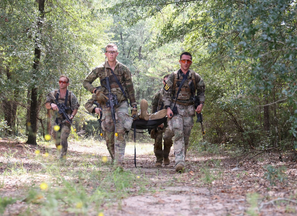 Four military personnel in camouflage uniforms are moving along a wooded dirt trail. Two soldiers in the center are carrying a person on a litter/stretcher, while two others flank them carrying rifles. The scene appears to depict a casualty evacuation (CASEVAC) drill or training exercise.