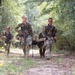 Four military personnel in camouflage uniforms are moving along a wooded dirt trail. Two soldiers in the center are carrying a person on a litter/stretcher, while two others flank them carrying rifles. The scene appears to depict a casualty evacuation (CASEVAC) drill or training exercise.
