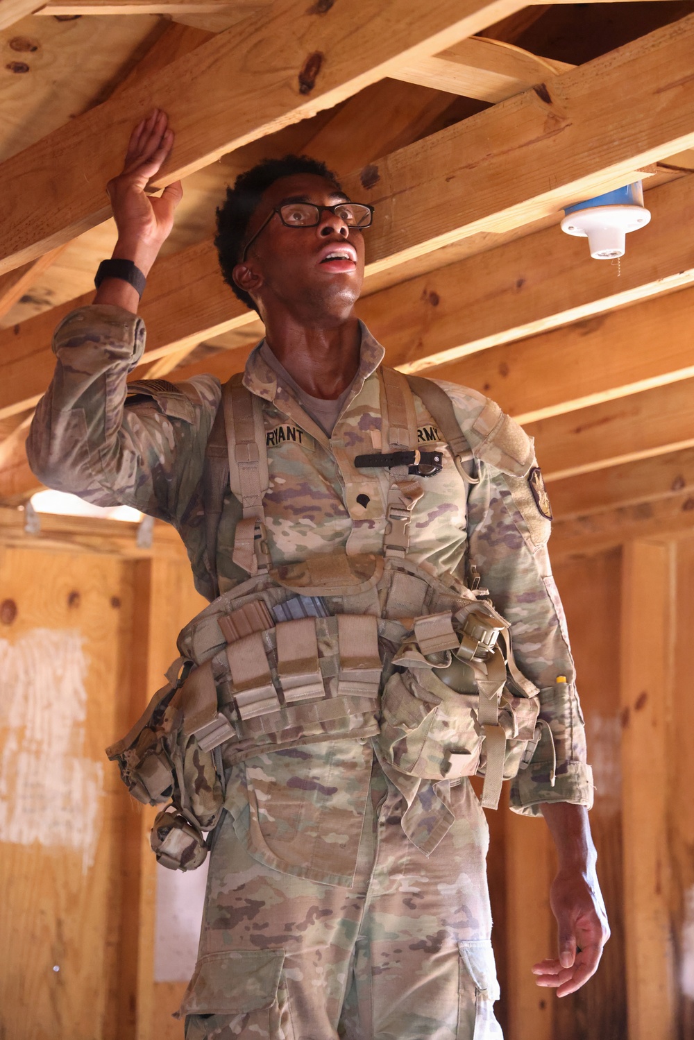 A U.S. Army soldier wearing Operational Camouflage Pattern (OCP) uniform and tactical vest is standing inside a wooden-framed structure, raising one hand toward the ceiling rafters while looking upward. The name tape visible on the uniform reads 'BRYANT' and the branch tape reads 'ARMY'. The soldier appears to be inspecting or pointing out something in the ceiling area of what appears to be a training or construction facility.