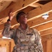 A U.S. Army soldier wearing Operational Camouflage Pattern (OCP) uniform and tactical vest is standing inside a wooden-framed structure, raising one hand toward the ceiling rafters while looking upward. The name tape visible on the uniform reads 'BRYANT' and the branch tape reads 'ARMY'. The soldier appears to be inspecting or pointing out something in the ceiling area of what appears to be a training or construction facility.