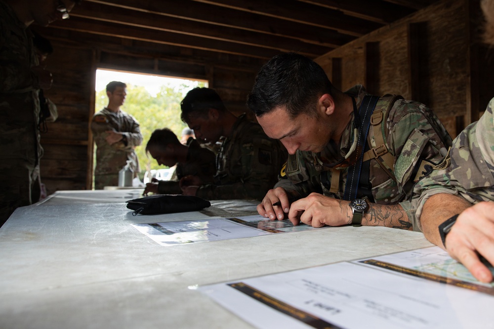 Military personnel in camouflage uniforms are gathered inside a wooden structure, studying maps and documents spread across a table. The scene appears to be a land navigation or mission planning exercise, with multiple soldiers focused on their materials. The primary subject in the foreground is closely examining a topographic or tactical map.