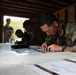 Military personnel in camouflage uniforms are gathered inside a wooden structure, studying maps and documents spread across a table. The scene appears to be a land navigation or mission planning exercise, with multiple soldiers focused on their materials. The primary subject in the foreground is closely examining a topographic or tactical map.