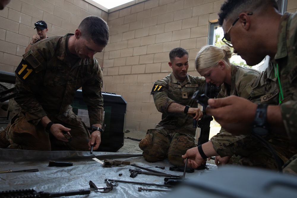 A group of U.S. Army soldiers in OCP (Operational Camouflage Pattern) uniforms are engaged in weapons maintenance or disassembly training. Multiple personnel are kneeling or sitting around a tarp on the ground covered with disassembled weapon components and tools. The setting appears to be an outdoor or semi-covered area adjacent to a concrete block wall.