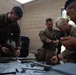A group of U.S. Army soldiers in OCP (Operational Camouflage Pattern) uniforms are engaged in weapons maintenance or disassembly training. Multiple personnel are kneeling or sitting around a tarp on the ground covered with disassembled weapon components and tools. The setting appears to be an outdoor or semi-covered area adjacent to a concrete block wall.