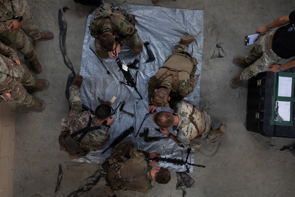 An overhead aerial view of military personnel in MultiCam/OCP camouflage uniforms conducting a weapons disassembly and reassembly training exercise. Several soldiers are gathered around a silver tarp on a concrete floor with disassembled rifles laid out. An observer or evaluator in dark clothing holds a clipboard on the right side, suggesting this is a formal assessment or timed competition.