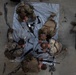An overhead aerial view of military personnel in MultiCam/OCP camouflage uniforms conducting a weapons disassembly and reassembly training exercise. Several soldiers are gathered around a silver tarp on a concrete floor with disassembled rifles laid out. An observer or evaluator in dark clothing holds a clipboard on the right side, suggesting this is a formal assessment or timed competition.
