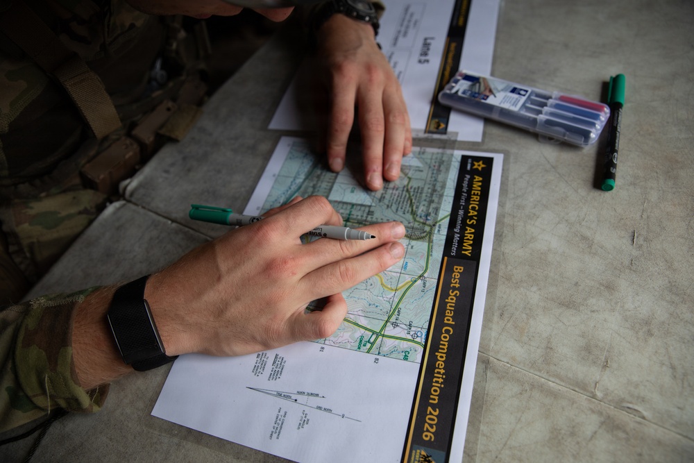 A soldier in camouflage uniform is working with a topographic map during America's Army Best Squad Competition 2026. The individual is holding markers and appears to be plotting or analyzing map coordinates. Multiple markers and reference documents are visible on the surface alongside the map.