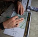 A soldier in camouflage uniform is working with a topographic map during America's Army Best Squad Competition 2026. The individual is holding markers and appears to be plotting or analyzing map coordinates. Multiple markers and reference documents are visible on the surface alongside the map.