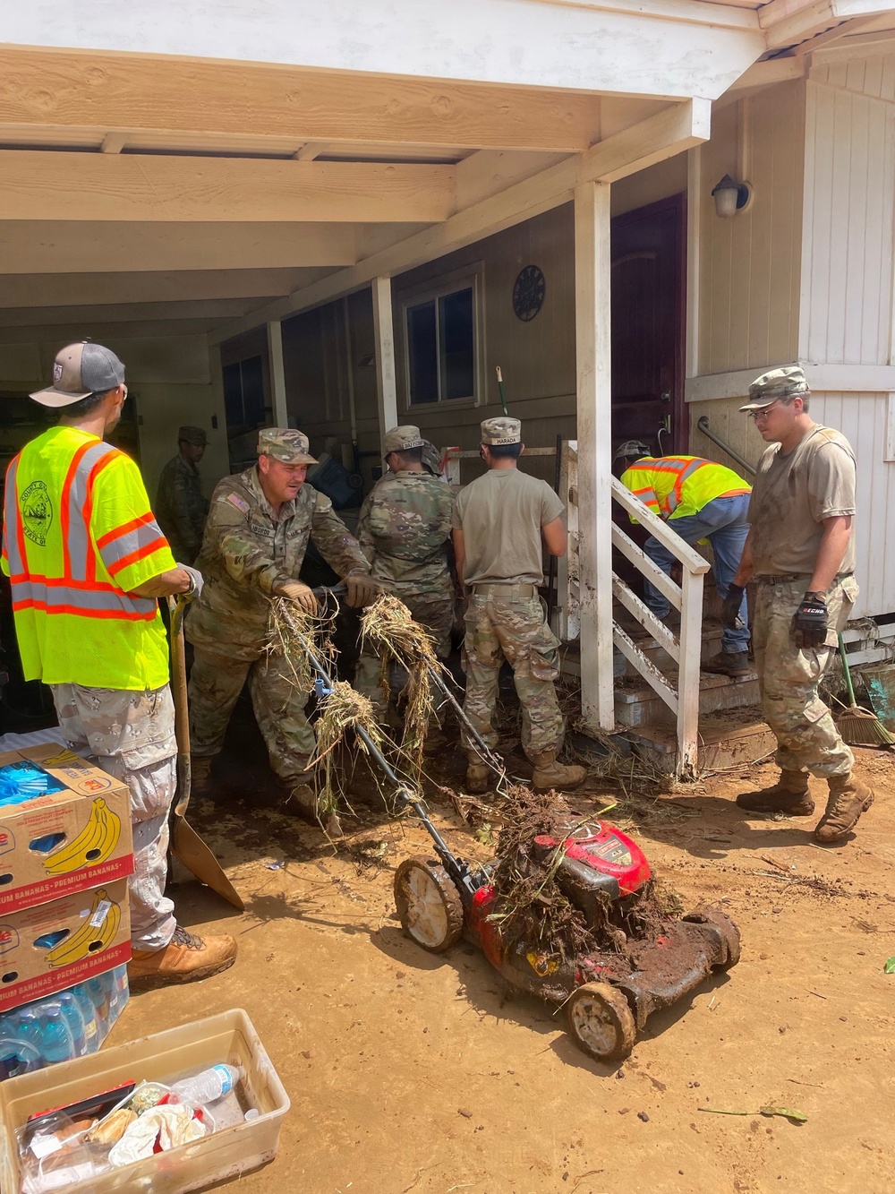 Military personnel in camouflage uniforms and civilian workers in high-visibility safety vests are conducting flood or disaster cleanup operations at a residential property. Workers are removing debris, mud-covered vegetation, and damaged items including a heavily mud-caked red lawn mower. The scene shows significant mud and debris accumulation consistent with flood damage recovery efforts.