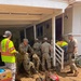 Military personnel in camouflage uniforms and civilian workers in high-visibility safety vests are conducting flood or disaster cleanup operations at a residential property. Workers are removing debris, mud-covered vegetation, and damaged items including a heavily mud-caked red lawn mower. The scene shows significant mud and debris accumulation consistent with flood damage recovery efforts.