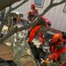 Military personnel in camouflage uniforms and high-visibility orange chainsaw safety gear are cutting up a large fallen tree near a building. At least three workers wearing orange helmets with face shields and hearing protection are actively using chainsaws, while a fourth person in standard uniform assists. The ground is covered in dark ash or charred debris, sawdust, and rocky material, suggesting post-fire disaster relief or debris clearing operations.