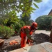 A worker in full personal protective equipment is using a chainsaw to cut through a large fallen tree trunk. The individual is wearing military camouflage clothing beneath bright orange high-visibility safety gear, including a Shindaiwa-branded chainsaw chaps, hard hat, and face shield. The setting appears to be a residential or landscaped area with visible debris, disturbed soil, and lush vegetation in the background.
