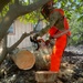 A worker in full personal protective equipment is actively cutting a large fallen tree trunk with a STIHL chainsaw. The worker is wearing camouflage clothing, bright orange high-visibility coveralls, a hard hat with face shield, and a face covering. Several freshly cut log sections are visible on the rocky ground, with a structure visible in the background.