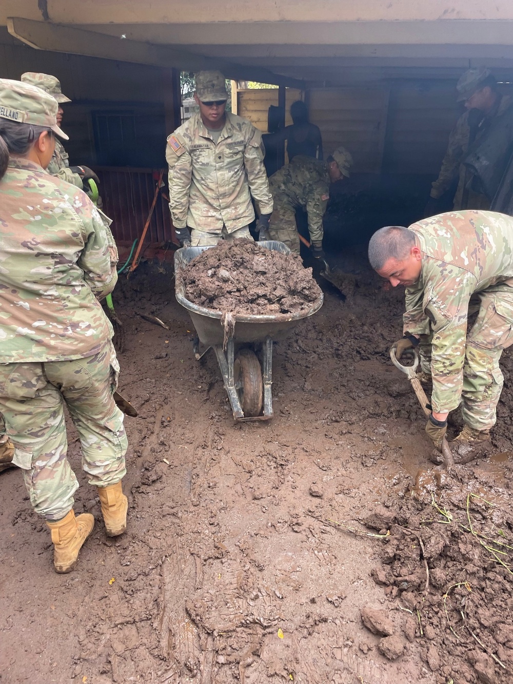 Multiple U.S. Army soldiers in OCP (Operational Camouflage Pattern) uniforms are engaged in flood or mudslide cleanup operations near a wooden structure. One soldier is pushing a wheelbarrow filled with mud while another shovels mud from the heavily saturated ground. The scene indicates disaster relief or emergency response work, with significant mud accumulation covering the entire ground surface.