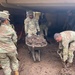 Multiple U.S. Army soldiers in OCP (Operational Camouflage Pattern) uniforms are engaged in flood or mudslide cleanup operations near a wooden structure. One soldier is pushing a wheelbarrow filled with mud while another shovels mud from the heavily saturated ground. The scene indicates disaster relief or emergency response work, with significant mud accumulation covering the entire ground surface.