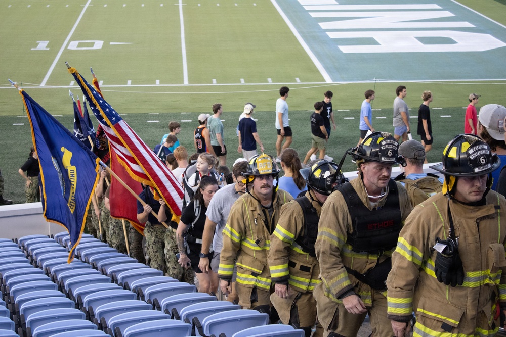 Firefighters in full turnout gear and helmets climb stadium steps alongside military personnel carrying flags, including the American flag and what appear to be branch-specific military flags. The event appears to be a memorial stair climb, likely a 9/11 tribute event held at a football stadium. Civilians and participants are also visible on the field and in the stands area.