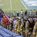 Firefighters in full turnout gear and helmets climb stadium steps alongside military personnel carrying flags, including the American flag and what appear to be branch-specific military flags. The event appears to be a memorial stair climb, likely a 9/11 tribute event held at a football stadium. Civilians and participants are also visible on the field and in the stands area.