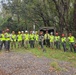 A group of approximately 13 military personnel and civilian workers pose for a group photo in a wooded outdoor area. They are equipped with safety gear including hard hats in red, yellow, and white, high-visibility vests, and gloves, along with chainsaws visible on the ground. The setting appears to be a disaster relief or debris clearing operation, with a military canvas-covered truck visible in the background.