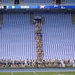 A large group of people, including military personnel in camouflage uniforms and civilians, are arranged in formation on the field and in the stands of a college football stadium identified as a Tar Heels venue. The scoreboard displays 'BALL ON 35', '2', and 'TAR HEELS' along with the number '25'. The stadium seats are largely empty, suggesting this is a pre-game ceremony or military appreciation event.