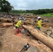 Workers in high-visibility safety vests and hard hats are conducting debris clearing operations in a flood or landslide-affected area. A worker is actively using a chainsaw to cut through a large fallen tree trunk amid scattered rocks, displaced soil, and debris. The landscape shows significant erosion and destruction, with green vegetation visible in the background suggesting a tropical or subtropical environment.