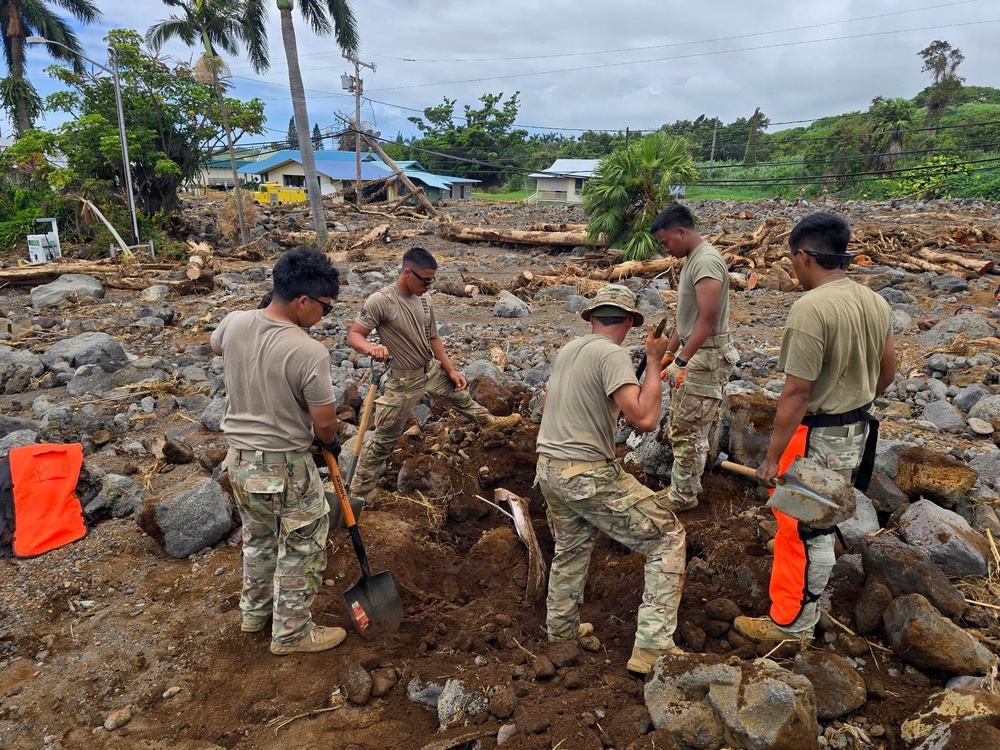 A group of military personnel in camouflage uniforms are conducting disaster recovery operations in a heavily debris-strewn area. The landscape shows extensive damage consistent with a flood or landslide event, with scattered rocks, uprooted trees, and destroyed vegetation across a wide area. Buildings and utility infrastructure are visible in the background amid the destruction.