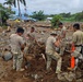 A group of military personnel in camouflage uniforms are conducting disaster recovery operations in a heavily debris-strewn area. The landscape shows extensive damage consistent with a flood or landslide event, with scattered rocks, uprooted trees, and destroyed vegetation across a wide area. Buildings and utility infrastructure are visible in the background amid the destruction.