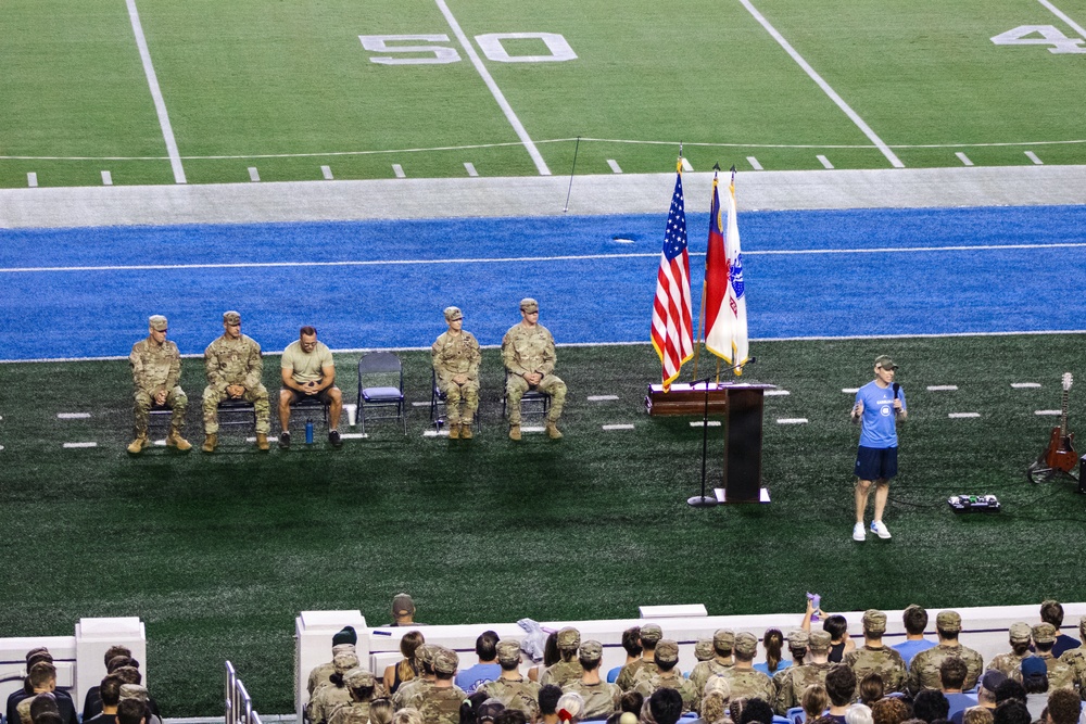 A military ceremony or event is taking place on an indoor football field, visible near the 50-yard line. Five uniformed military personnel in camouflage (ACU/OCP) are seated in folding chairs on the field, while a civilian in a blue shirt speaks at a podium flanked by flags. A large audience, consisting of both military personnel and civilians, observes from the stands.