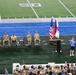 A military ceremony or event is taking place on an indoor football field, visible near the 50-yard line. Five uniformed military personnel in camouflage (ACU/OCP) are seated in folding chairs on the field, while a civilian in a blue shirt speaks at a podium flanked by flags. A large audience, consisting of both military personnel and civilians, observes from the stands.