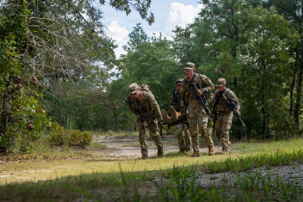 A group of military personnel in full combat gear are conducting a casualty evacuation drill in a wooded training area. Three soldiers are carrying what appears to be a simulated casualty on a litter or improvised stretcher along a dirt path. The setting appears to be a military training installation with mixed pine and deciduous forest.