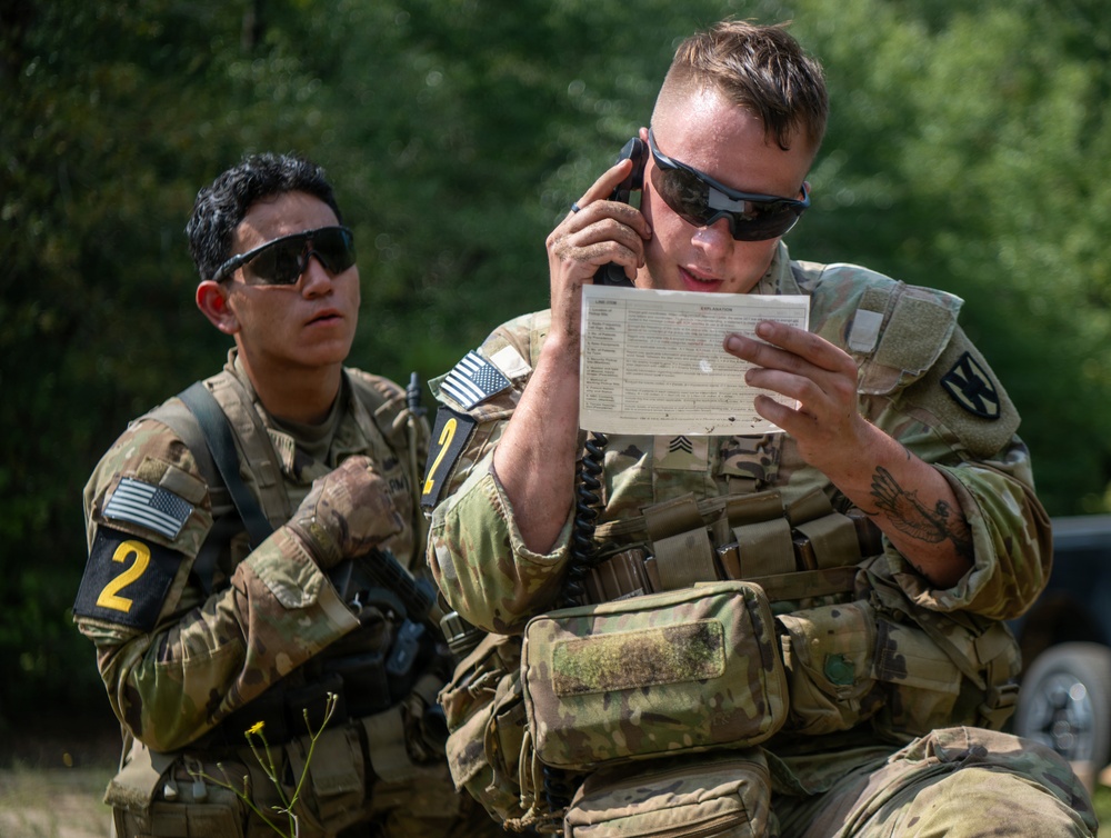 Two U.S. Army soldiers in full combat gear are engaged in what appears to be a field training exercise. The soldier in the foreground is speaking on a radio handset while reading from a reference card, likely a call-for-fire or MEDEVAC request card. Both soldiers wear OCP (Operational Camouflage Pattern) uniforms with American flag patches and numbered armbands indicating an exercise or competition setting.