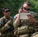 Two U.S. Army soldiers in full combat gear are engaged in what appears to be a field training exercise. The soldier in the foreground is speaking on a radio handset while reading from a reference card, likely a call-for-fire or MEDEVAC request card. Both soldiers wear OCP (Operational Camouflage Pattern) uniforms with American flag patches and numbered armbands indicating an exercise or competition setting.