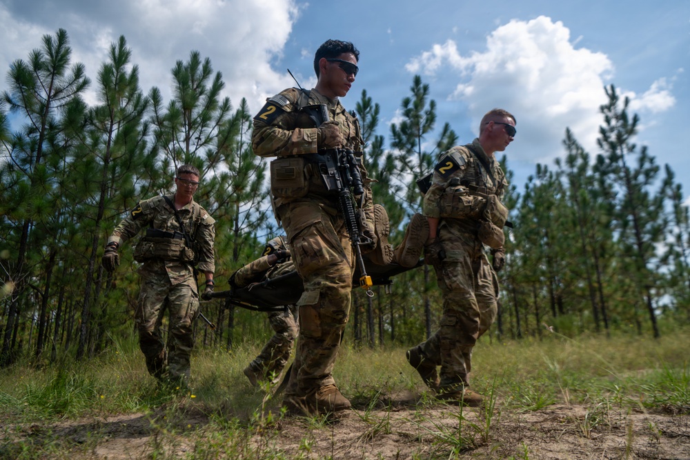 Three soldiers in U.S. Army camouflage uniforms are carrying a litter (stretcher) across an open sandy trail bordered by pine trees, suggesting a field training exercise or casualty evacuation drill. Two of the visible soldiers wear the number '2' on their uniforms, indicating team or squad designation. The setting appears to be a military training area in a southeastern U.S. pine forest environment.