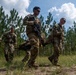 Three soldiers in U.S. Army camouflage uniforms are carrying a litter (stretcher) across an open sandy trail bordered by pine trees, suggesting a field training exercise or casualty evacuation drill. Two of the visible soldiers wear the number '2' on their uniforms, indicating team or squad designation. The setting appears to be a military training area in a southeastern U.S. pine forest environment.