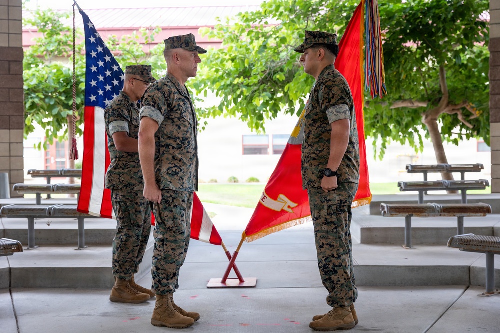 Three U.S. Marines in woodland MARPAT camouflage uniforms are participating in a formal military ceremony, likely a change of command or similar official event. Two crossed flags — an American flag and a Marine Corps unit guidon — are displayed on a stand between the central figures. The setting appears to be a covered outdoor pavilion at a military installation.