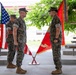 Three U.S. Marines in woodland MARPAT camouflage uniforms are participating in a formal military ceremony, likely a change of command or similar official event. Two crossed flags — an American flag and a Marine Corps unit guidon — are displayed on a stand between the central figures. The setting appears to be a covered outdoor pavilion at a military installation.