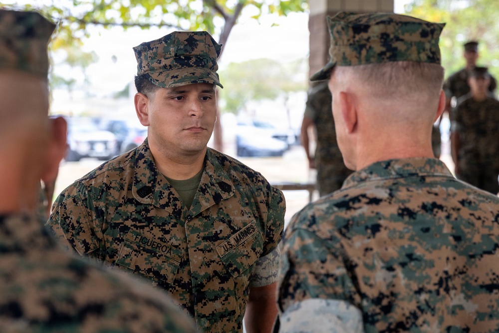 A group of U.S. Marines in MARPAT woodland camouflage uniforms are engaged in a face-to-face conversation or briefing in an outdoor covered area. The central figure, identified by his name tape as 'FIGUEROA,' appears to be speaking with or listening to a senior Marine whose back is to the camera. The setting appears to be a military installation, with vehicles and trees visible in the background.