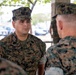 A group of U.S. Marines in MARPAT woodland camouflage uniforms are engaged in a face-to-face conversation or briefing in an outdoor covered area. The central figure, identified by his name tape as 'FIGUEROA,' appears to be speaking with or listening to a senior Marine whose back is to the camera. The setting appears to be a military installation, with vehicles and trees visible in the background.