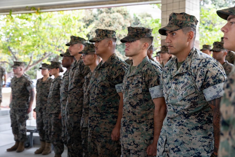 A formation of U.S. Marines standing at attention in MARPAT (Marine Pattern) digital camouflage uniforms. The personnel are arranged in rows, appearing to be participating in a formal military ceremony or formation event. The setting appears to be an outdoor covered area with trees visible in the background.