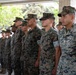 A formation of U.S. Marines standing at attention in MARPAT (Marine Pattern) digital camouflage uniforms. The personnel are arranged in rows, appearing to be participating in a formal military ceremony or formation event. The setting appears to be an outdoor covered area with trees visible in the background.