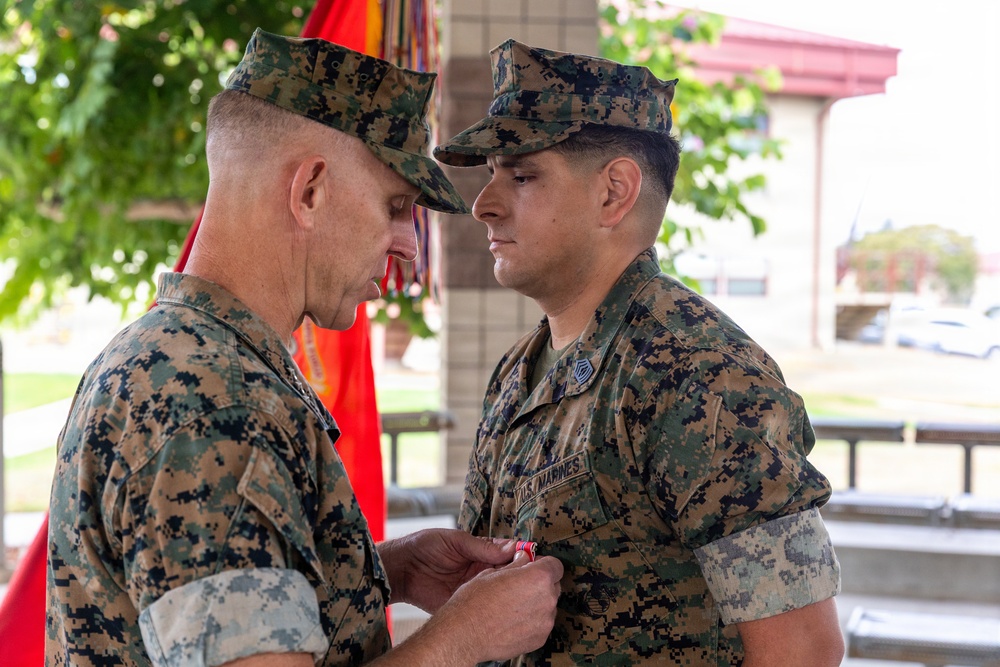Two U.S. Marines in MARPAT woodland camouflage uniforms are engaged in a military awards ceremony. A senior Marine is pinning a medal or decoration onto the chest of a younger Marine. A red Marine Corps flag is visible in the background, indicating a formal military occasion.