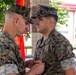 Two U.S. Marines in MARPAT woodland camouflage uniforms are engaged in a military awards ceremony. A senior Marine is pinning a medal or decoration onto the chest of a younger Marine. A red Marine Corps flag is visible in the background, indicating a formal military occasion.