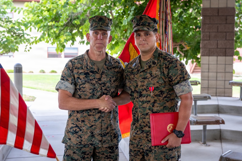 Two U.S. Marines in MARPAT woodland camouflage uniforms are shaking hands in what appears to be a military award ceremony. The Marine on the right is wearing a medal on his chest and holding a red presentation folder, indicating he has just received a commendation or decoration. A U.S. Marine Corps flag and American flag are visible in the background.