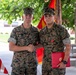 Two U.S. Marines in MARPAT woodland camouflage uniforms are shaking hands in what appears to be a military award ceremony. The Marine on the right is wearing a medal on his chest and holding a red presentation folder, indicating he has just received a commendation or decoration. A U.S. Marine Corps flag and American flag are visible in the background.